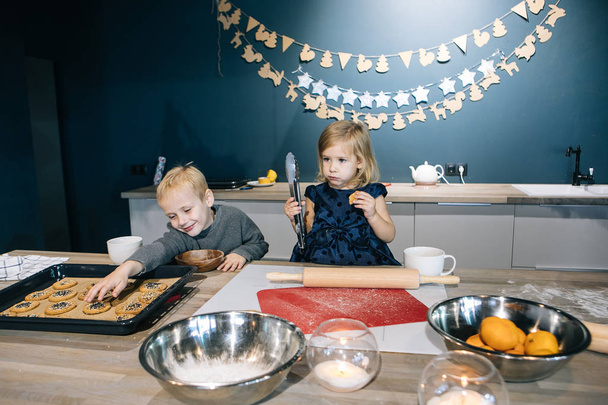 Blond children - a boy and a girl - are preparing cookies and a cake for Christmas in the kitchen, flour, rolling pin, and children's emotions - Фото, зображення