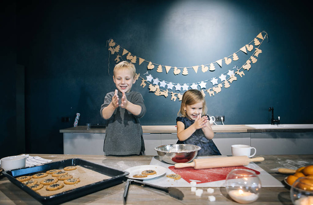 Blond children - a boy and a girl - are preparing cookies and a cake for Christmas in the kitchen, flour, rolling pin, and children's emotions - Zdjęcie, obraz