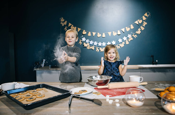 Blond children - a boy and a girl - are preparing cookies and a cake for Christmas in the kitchen, flour, rolling pin, and children's emotions - Zdjęcie, obraz