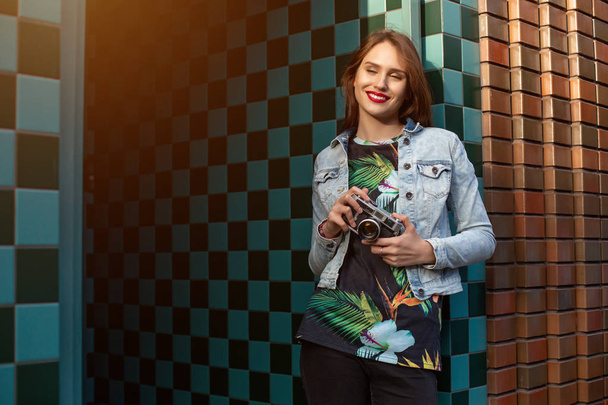 Крутая смешная девушка модель с ретро-кинокамерой в джинсовой куртке, темные волосы на открытом воздухе над городской стеной на фоне клетки. Солнечная вспышка
 - Фото, изображение