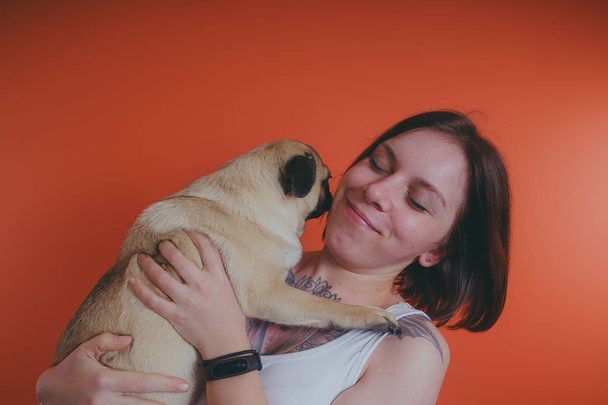Beautiful girl with tattoo holding cute pug puppy on her hands, on orange background. The concept of friendship between man and dog. - 写真・画像