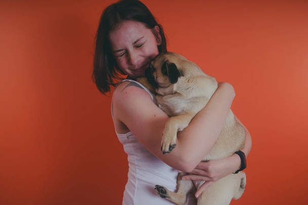 Beautiful girl with tattoo holding cute pug puppy on her hands, on orange background. The concept of friendship between man and dog. - Foto, afbeelding