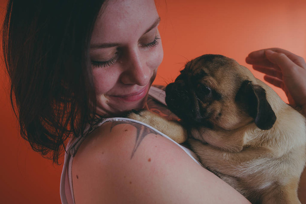 Beautiful girl with tattoo holding cute pug puppy on her hands, on orange background. The concept of friendship between man and dog. - Фото, изображение