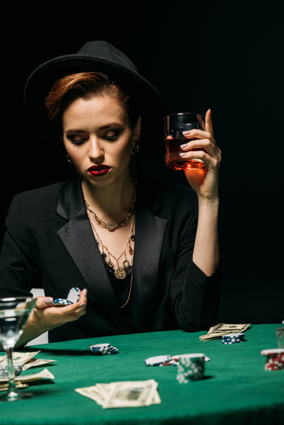 beautiful girl in jacket and hat holding glass of whiskey and poker chips at table in casino - Foto, Bild