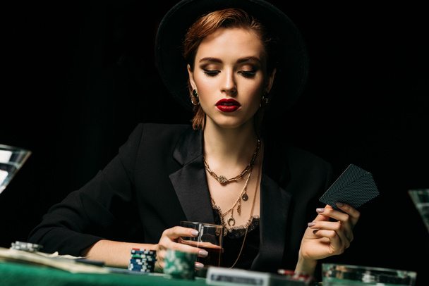 surface level of attractive girl in jacket and hat holding glass of whiskey and playing poker in casino - Photo, Image