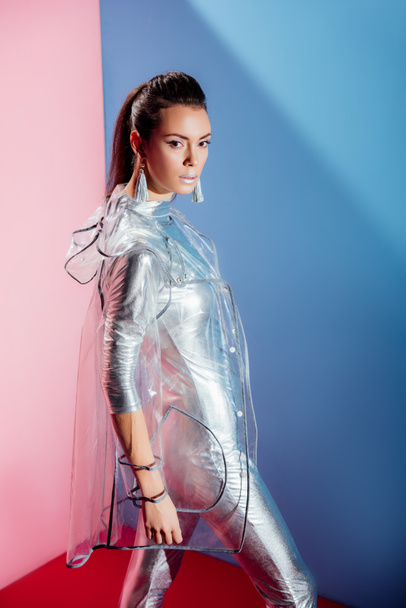fashionable young woman posing in silver bodysuit and raincoat on pink and blue background - Foto, Imagen