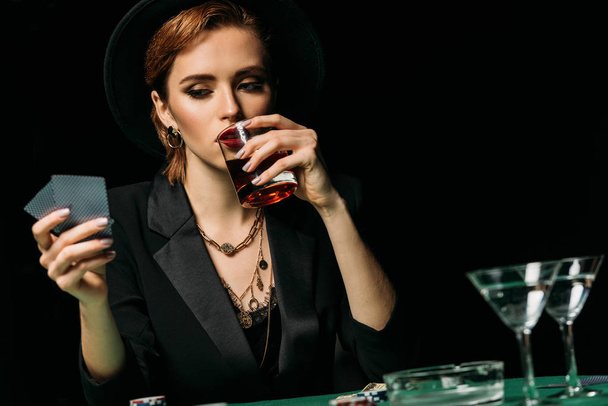 attractive girl in jacket and hat drinking whiskey and looking at poker cards in casino - Фото, изображение