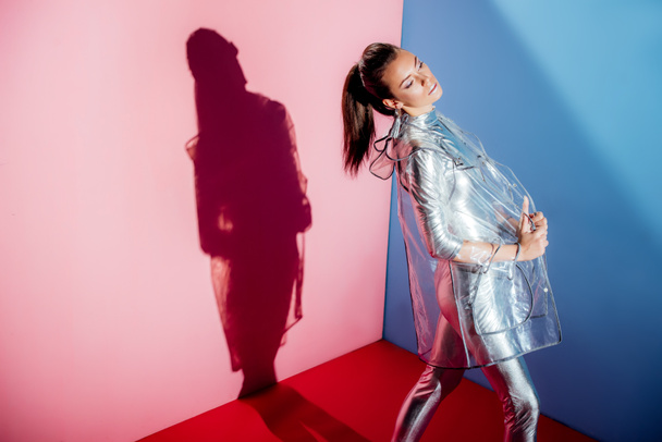 elegant girl posing in metallic bodysuit and trendy raincoat for fashion shoot on pink and blue background - Photo, Image