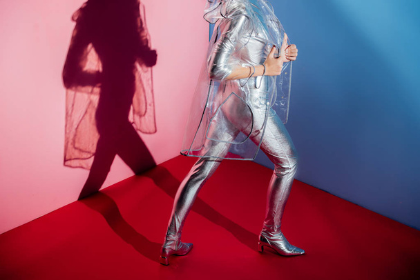 cropped view of fashionable girl posing in silver bodysuit and raincoat on pink and blue background - Fotografie, Obrázek