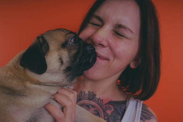 Beautiful girl with tattoo holding cute pug puppy on her hands, on orange background. The concept of friendship between man and dog. - Foto, Bild