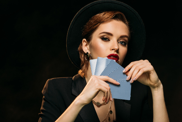 beautiful girl in jacket and hat covering face with poker cards and looking at camera isolated on black - Photo, Image
