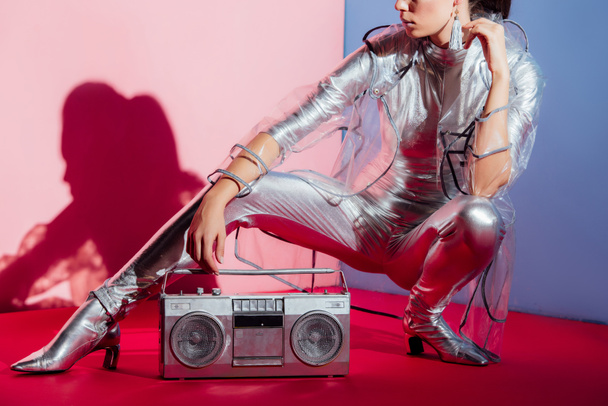 cropped view of model in silver bodysuit and raincoat posing with retro boombox on pink and blue background - Fotó, kép
