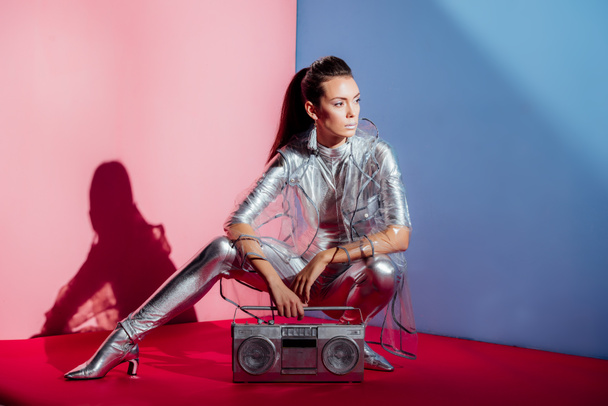 fashionable model in silver bodysuit and raincoat posing with boombox on pink and blue background - Photo, Image