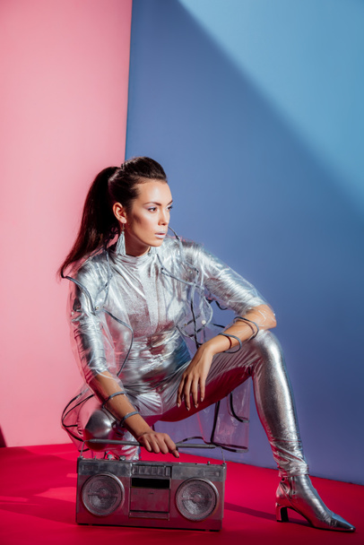 fashionable young woman in silver bodysuit and raincoat posing with boombox on pink and blue background - Fotoğraf, Görsel