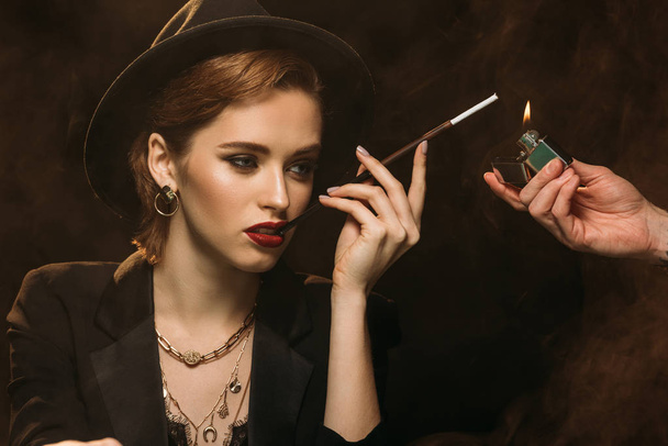 man lighting cigarette to attractive girl in jacket and hat on black - Фото, зображення
