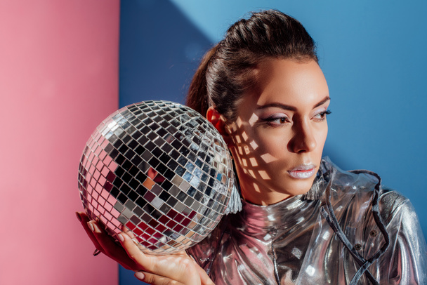 beautiful stylish young woman posing with silver disco ball on pink and blue background - Фото, изображение