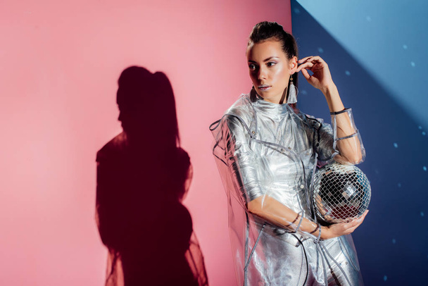 stylish young woman in metallic bodysuit and raincoat posing with disco ball on pink and blue background - Foto, Bild