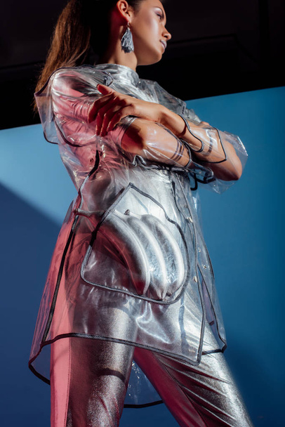 bottom view of fashionable model in metallic bodysuit and raincoat posing with silver bananas on blue background - Photo, Image