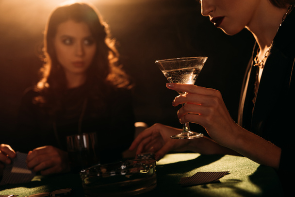カジノ、カクテルのガラスを保持している女性でのテーブルでポーカーをプレイする魅力的な女の子のトリミングされた画像 - 写真・画像