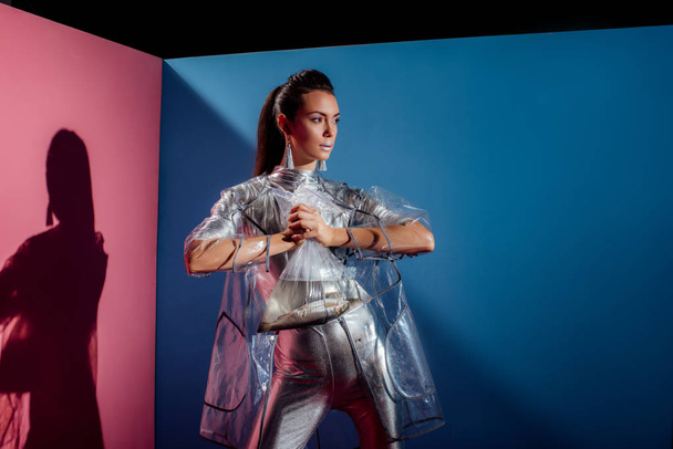 trendy young woman in silver bodysuit and raincoat holding plastic bag with fish for fashion shoot on pink and blue background - Photo, Image