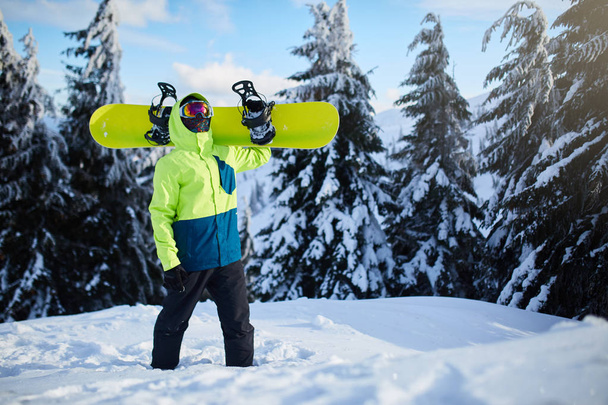 Snowboarder climbing to mountain top carrying his snowboard through forest for backcountry freeride and wearing reflective goggles, colorful fashion outfit and balaclava at ski resort. Winter sports. - Φωτογραφία, εικόνα