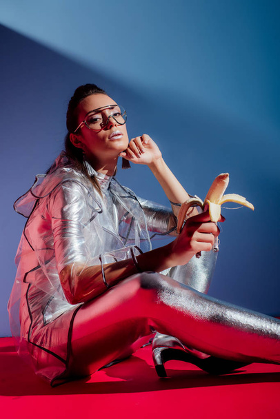 attractive fashionable girl in metallic bodysuit and raincoat posing with silver banana on pink and blue background - Foto, immagini