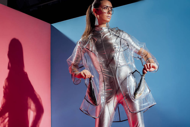 beautiful stylish girl in silver bodysuit and raincoat posing with fish on pink and blue background - Fotó, kép