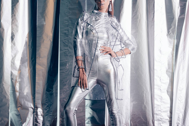 cropped view of young fashionable woman posing in silver bodysuit and raincoat on metallic background - Фото, зображення