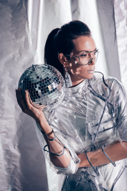 beautiful stylish young woman in silver bodysuit and raincoat posing with disco ball on metallic background - Foto, Imagen