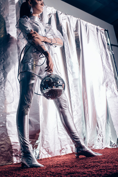 bottom view of fashionable girl in silver bodysuit and raincoat posing with disco ball on metallic background - Φωτογραφία, εικόνα
