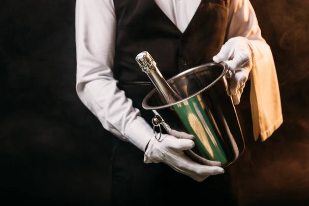 cropped image of waiter holding bottle of alcohol champagne in bucket on black - Foto, Imagen