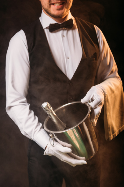 cropped image of smiling waiter holding bottle of champagne in bucket on black - Foto, Bild