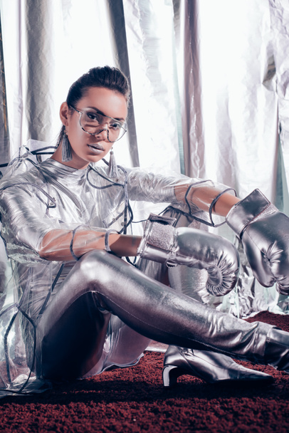 fashionable model posing in bodysuit, raincoat and silver box gloves on metallic background - Valokuva, kuva