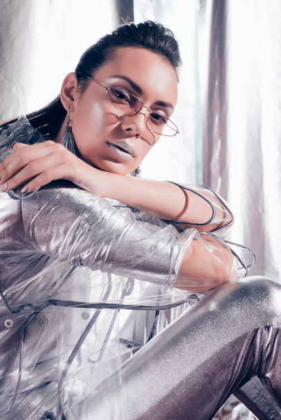 fashionable girl posing in trendy glasses, silver bodysuit and raincoat on metallic background - Foto, afbeelding