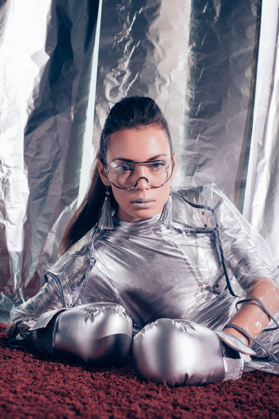 attractive fashionable girl posing in bodysuit, raincoat and silver box gloves on metallic background - Photo, Image