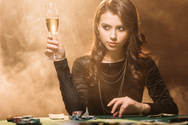 beautiful woman holding glass of champagne at poker table in casino and looking away - Photo, Image