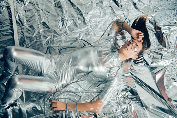 top view of stylish woman posing in silver bodysuit and raincoat on metallic background - Fotó, kép