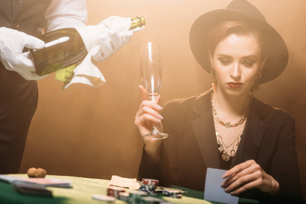 waiter pouring champagne in glass of attractive girl in jacket and hat in casino, she playing poker - Photo, Image