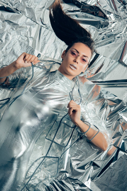 top view of brunette fashionable girl posing in silver bodysuit and raincoat on metallic background - Photo, Image