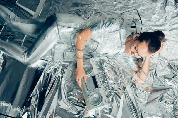 top view of attractive woman in silver bodysuit and raincoat posing with boombox on metallic background - Foto, afbeelding