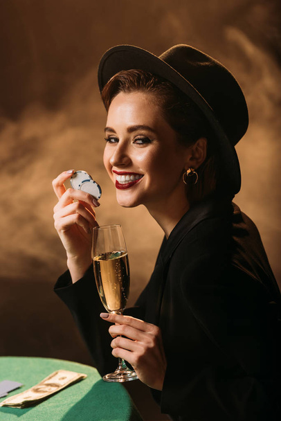 happy attractive girl in jacket and hat holding glass of champagne and poker chips at table in casino, looking at camera - Foto, Bild