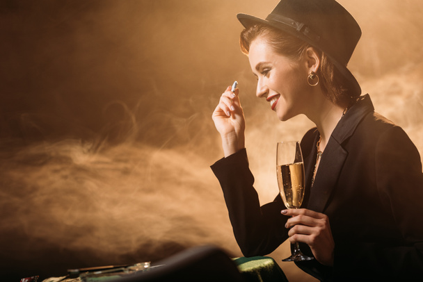 Zijaanzicht van lachende aantrekkelijk meisje in jas en hoed bedrijf glas champagne en poker chips aan tafel in casino - Foto, afbeelding