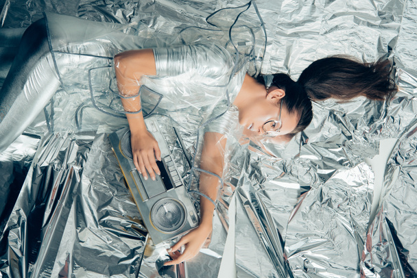 top view of elegant woman in silver bodysuit and raincoat posing with retro boombox on metallic background - Фото, изображение
