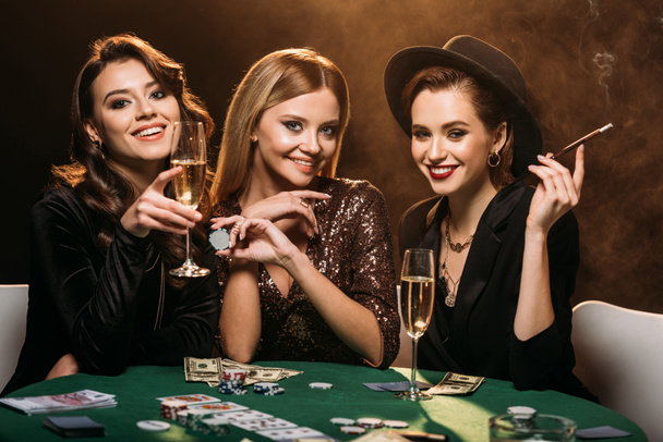 Ελκυστικές κοπέλες με ποτήρι σαμπάνια, τσιγάρο και πόκερ μάρκες κάθεται σε τραπέζι σε καζίνο και βλέπουν τα φωτογραφικών μηχανών - Φωτογραφία, εικόνα