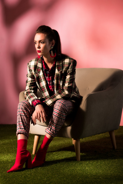 beautiful young woman in checkered suit posing in armchair on pink background - Photo, Image