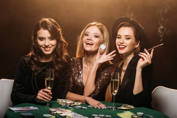 Смеющиеся привлекательные девушки с бокалом шампанского, сигаретами и фишками для покера, сидящие за столом в казино
 - Фото, изображение