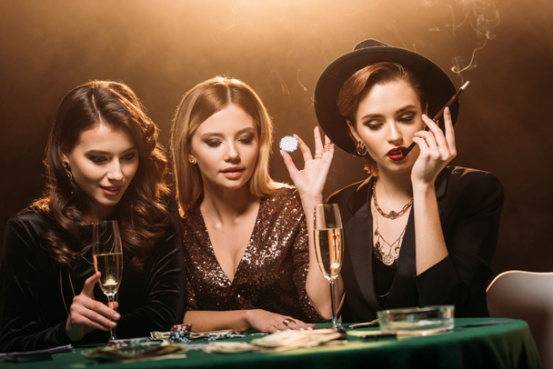 attractive girls with glass of champagne, cigarette and poker chips sitting at table and looking at poker cards in casino - Photo, Image