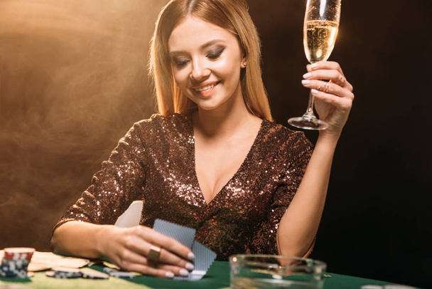 smiling attractive girl holding glass of champagne and looking at poker cards at table in casino - Fotó, kép