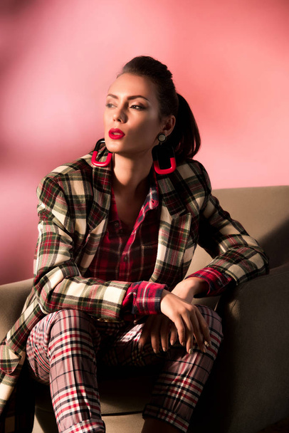beautiful stylish girl in checkered suit posing in armchair on pink background - Фото, изображение