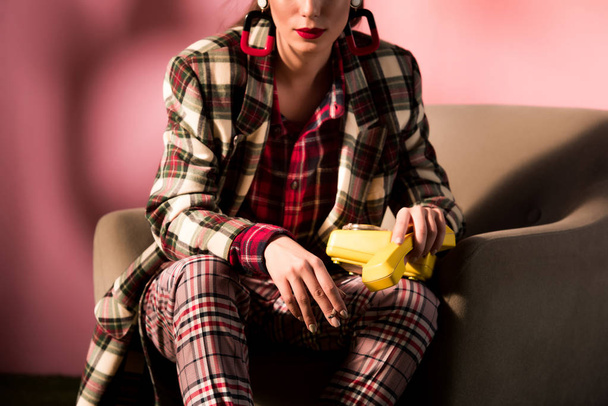 cropped view of elegant girl in checkered suit posing in armchair with retro telephone on pink background - Фото, зображення